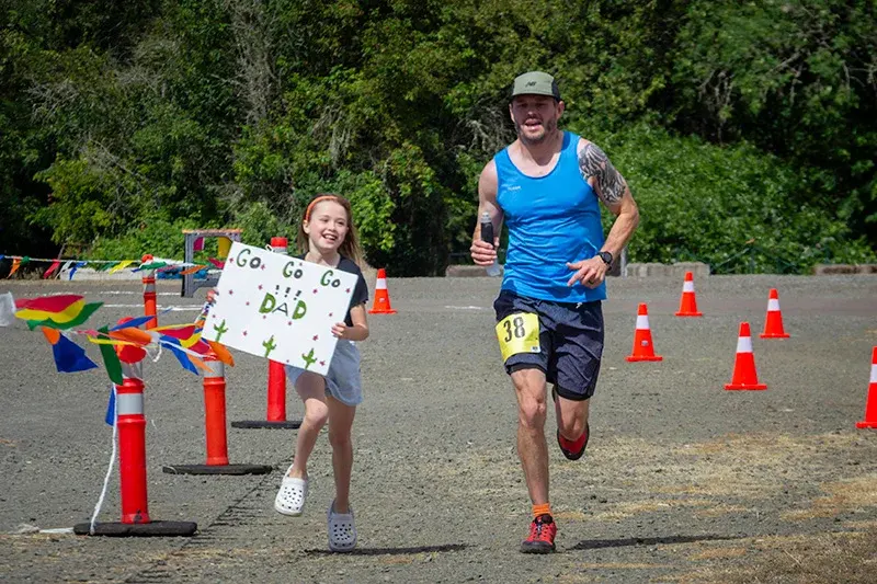 Runner crosses finish line with daughter cheering alongside holding a 'Go Go Go Dad' sign